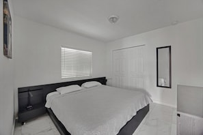 A bedroom with a bed, a bedside table, a mirror, a window with blinds, and a door.