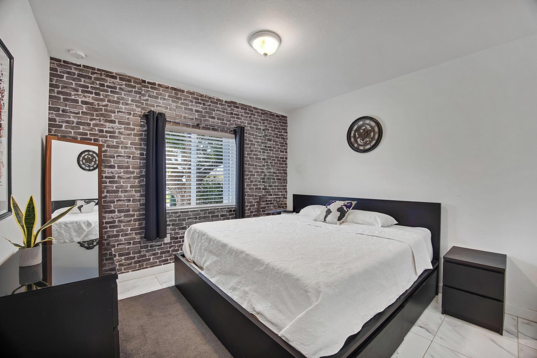 A bedroom with a brick wall, a bed with white bedding, a bedside table, a nightstand, a mirror, and a clock on the wall.