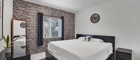 A bedroom with a brick wall, a bed with white bedding, a bedside table, a nightstand, a mirror, and a clock on the wall.