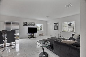 A modern living room with a flat-screen TV, a glass coffee table, a sofa, and a dining area with chairs. - Close To Airport, Miami & Wynwood New Modern Home! (Miami)