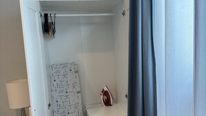 A room with a white wardrobe, a small ironing board with an iron, and a lamp on a bedside table.