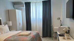 A modern hotel room with a neatly made bed, a desk with a computer and remote controls, and a window with curtains.