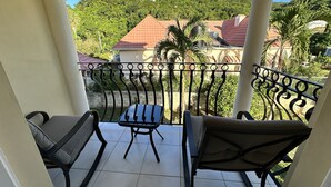 A balcony with two chairs and a small table, overlooking a green hillside with houses. - Villa Nilaja (Montego Bay)