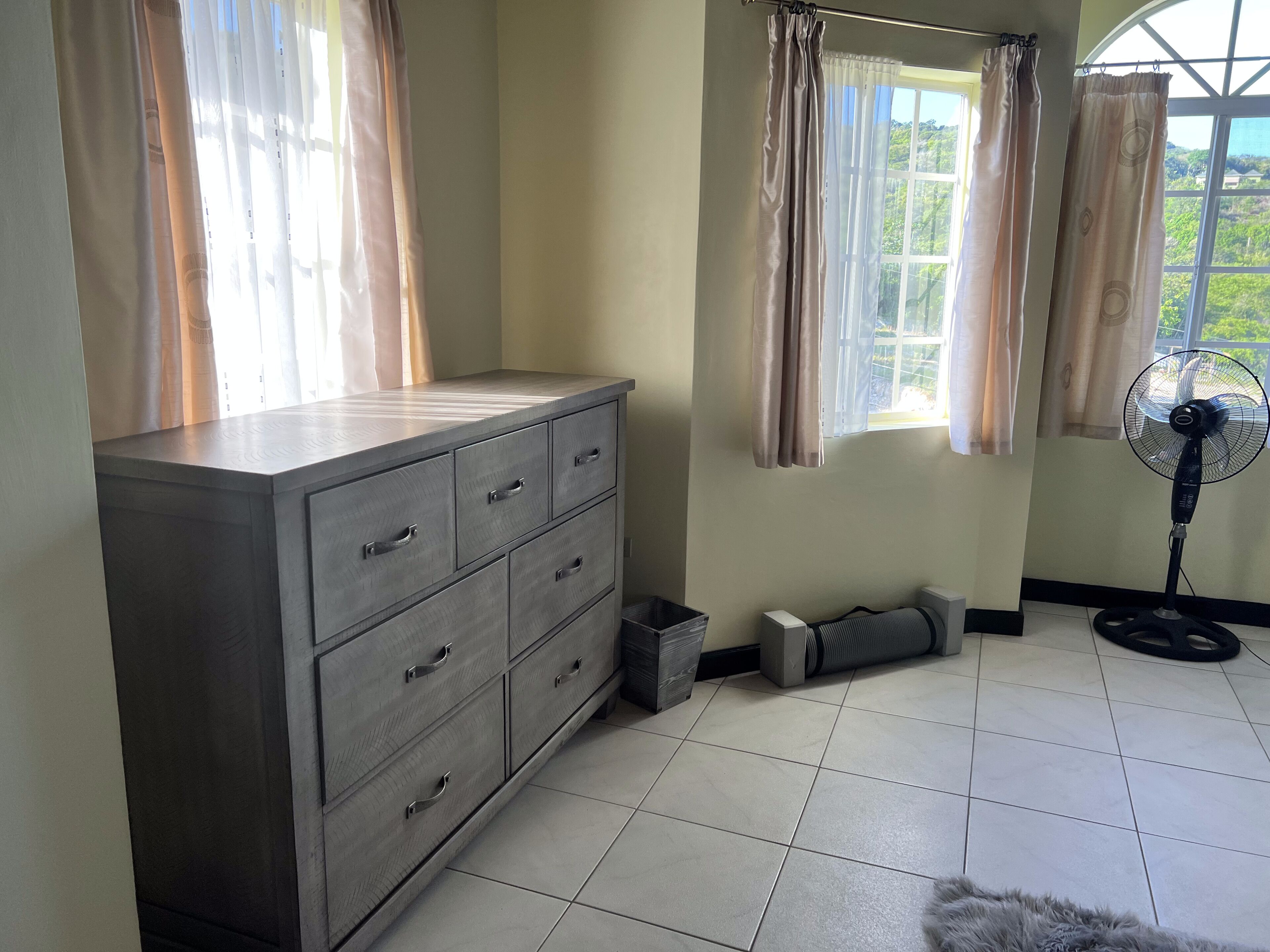 Une pièce avec une grande commode en bois, un ventilateur sur pied et une fenêtre avec des rideaux.