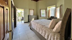 A bedroom with a bed, a dresser, a chair, and a fan. There is a door leading to a balcony with a view of palm trees. - Villa Nilaja (Montego Bay)