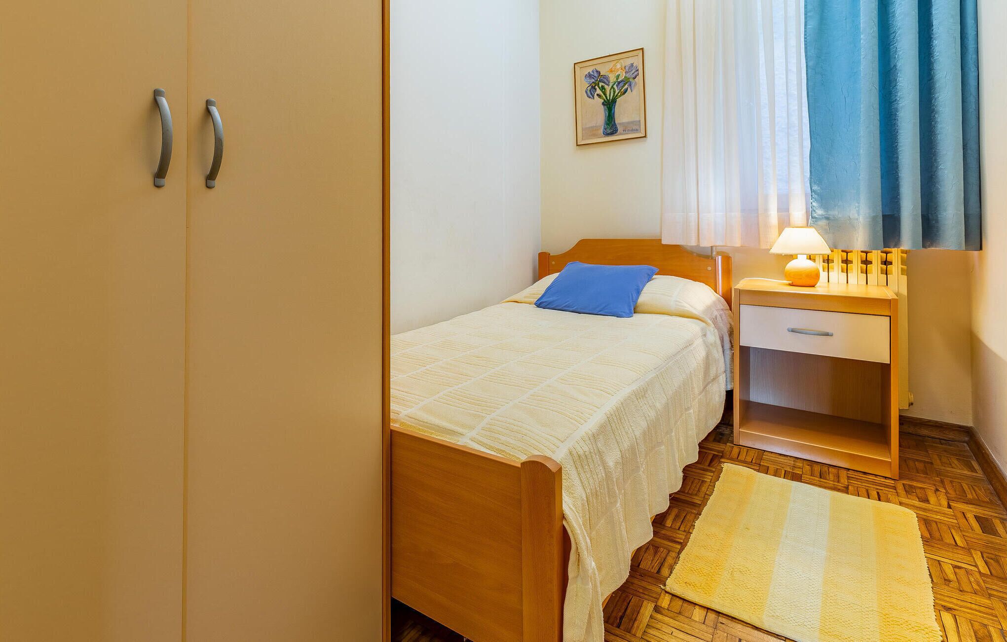 A bedroom with a wooden bed, a nightstand with a lamp, a blue pillow, a painting on the wall, and a wooden floor.