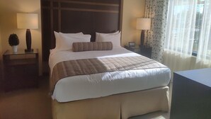 Ein Hotelzimmer mit einem großen Bett, zwei Nachttischen, einer Lampe, einem Topf mit einer Pflanze und einem Fenster mit Vorhängen.