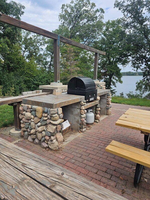 Un área de parrilla al aire libre con un muro de piedra, una mesa de madera y vistas a un lago.