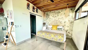 A bedroom with a bed, a ceiling fan, a wooden ceiling, a wall with a geometric pattern, and a window.