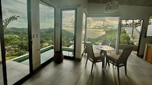 A spacious room with a large glass door leading to a balcony with a view of a lush landscape. There is a round table with chairs in the center of the room.