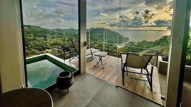 A balcony with a glass table and chairs, overlooking a scenic landscape with hills and a body of water.