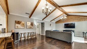 Een ruime woonkamer met een houten plafond, een grote grijze bank, houten krukken, een kroonluchter en een flatscreen televisie die aan de muur is bevestigd.