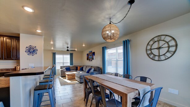 Een ruime keuken met een eethoek, met een houten tafel en blauwe stoelen, een grote klok aan de muur en een plafondventilator.