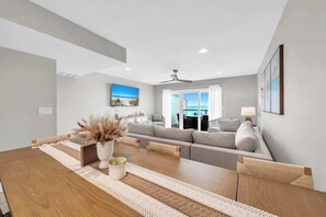 Uma sala de estar moderna com uma mesa de jantar de madeira, sofá cinza e vista para o oceano através de grandes janelas.