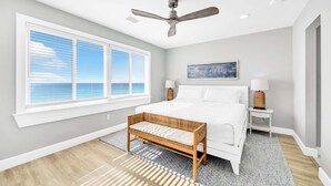A bedroom with a large window, a bed, a bench, a ceiling fan, and a wall-mounted lamp. - Summerhaven 3 | Beach Front Townhome with Free Chairs through October! | New Coastal Design! | Tail-Wagging Approved (Miramar Beach)