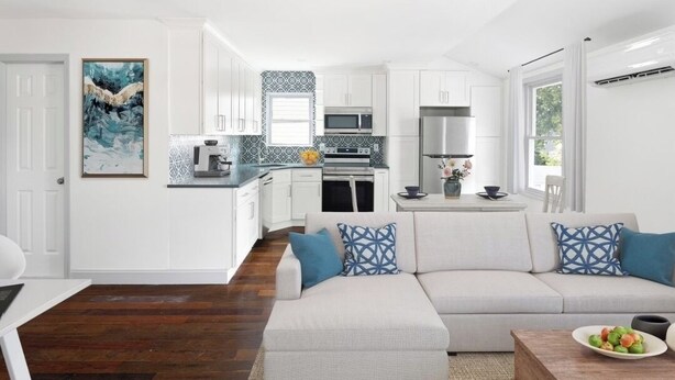 A modern kitchen with white cabinets, a microwave, and a stove. There is a dining area with a white table and a sofa with patterned cushions.