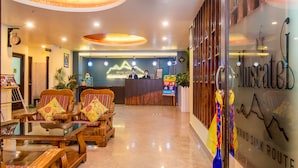 A hotel lobby with a reception desk, two wooden armchairs with colorful cushions, a glass table, and a wall with the hotel's name and a mountain graphic. - Muscatel Grand Silkroute 300 Mts MG Marg (Gangtok)