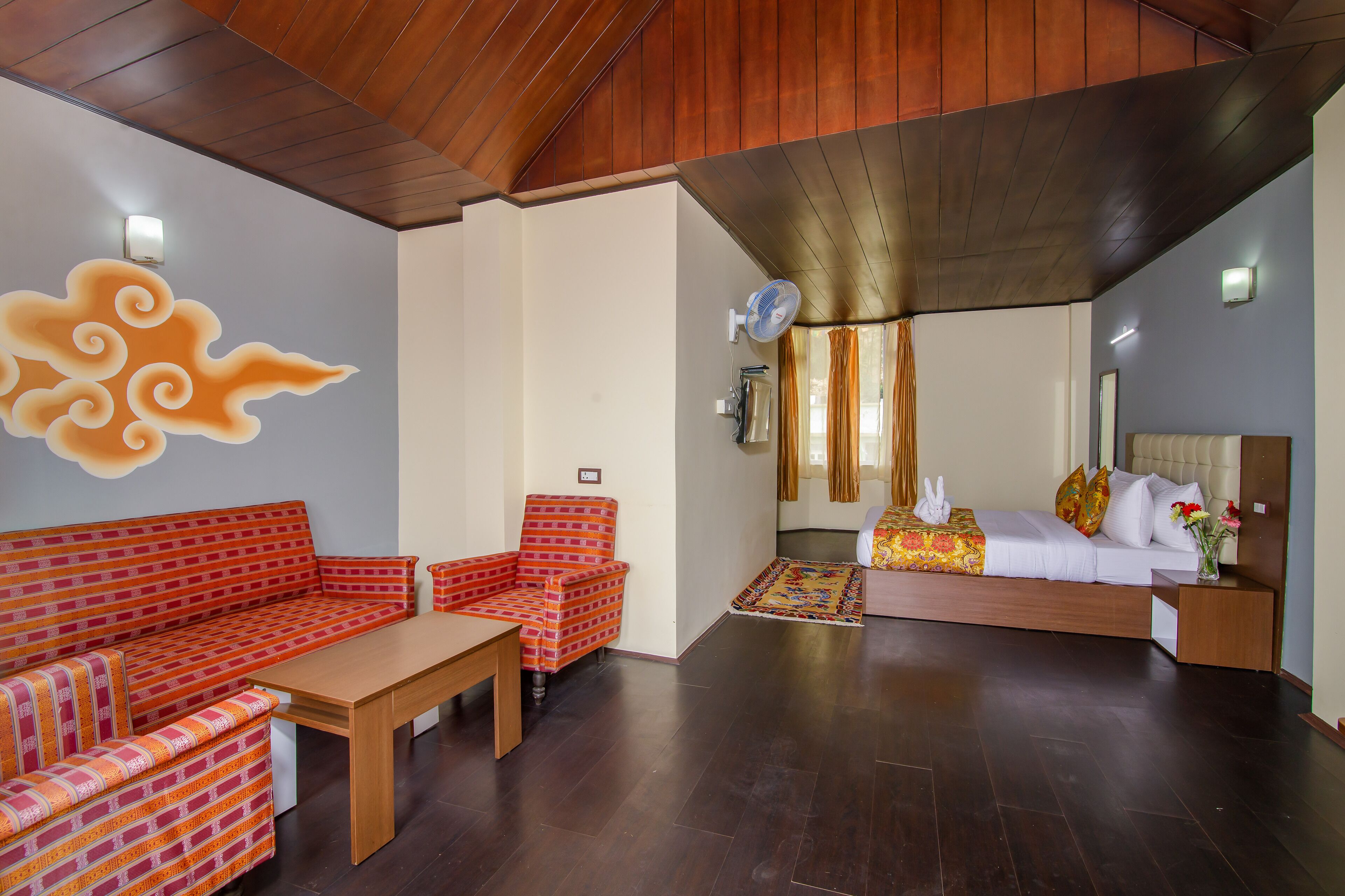 A room with a wooden ceiling, a sofa, a chair, a bed with a white headboard, and a small table.