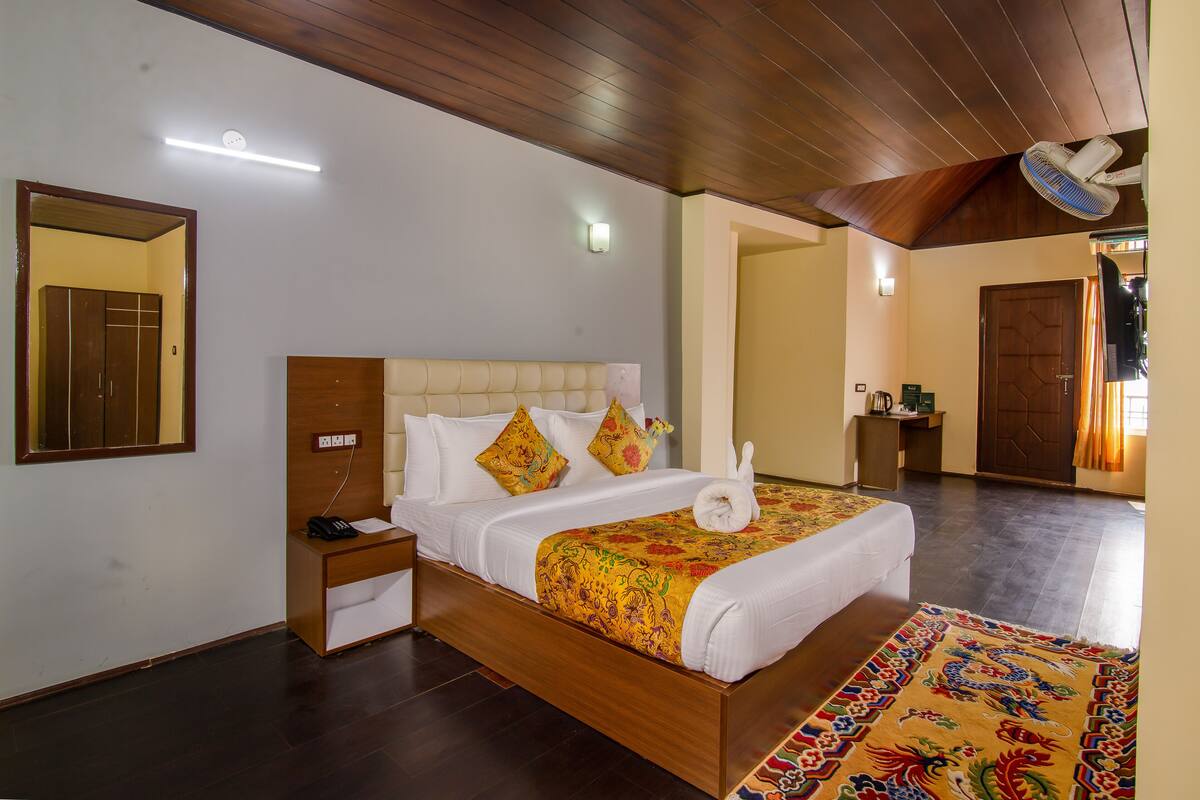 Une chambre d’hôtel avec un lit en bois, des tables de chevet, une petite table, une lampe fixée au mur et un ventilateur de plafond.
