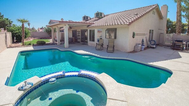 A swimming pool with a hot tub in the foreground, a covered patio area with chairs, and a house with a tiled roof in the background.