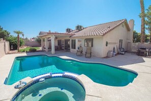 Una piscina con un jacuzzi en primer plano, un área de patio techada con sillas, y una casa con techo de tejas al fondo.