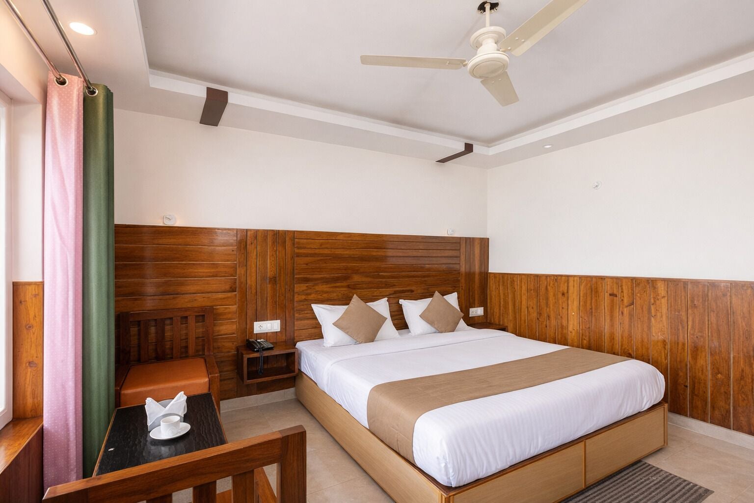 Habitación de hotel con cabecera de madera, cama con ropa de cama blanca, mesita de noche con lámpara, un pequeño banco de madera y un ventilador de techo.