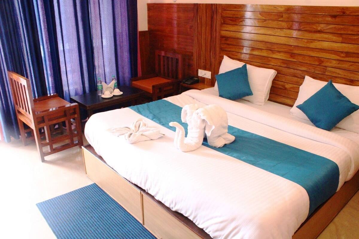 A hotel room with a wooden headboard, a bed with white and teal bedding, a wooden chair, a small table with a bottle and glasses, and a blue rug.