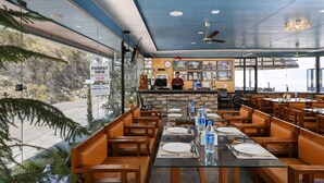 Un restaurant offrant une vue sur une forêt, avec des tables et des bancs en bois, un comptoir où se trouve un employé, et une horloge au mur.