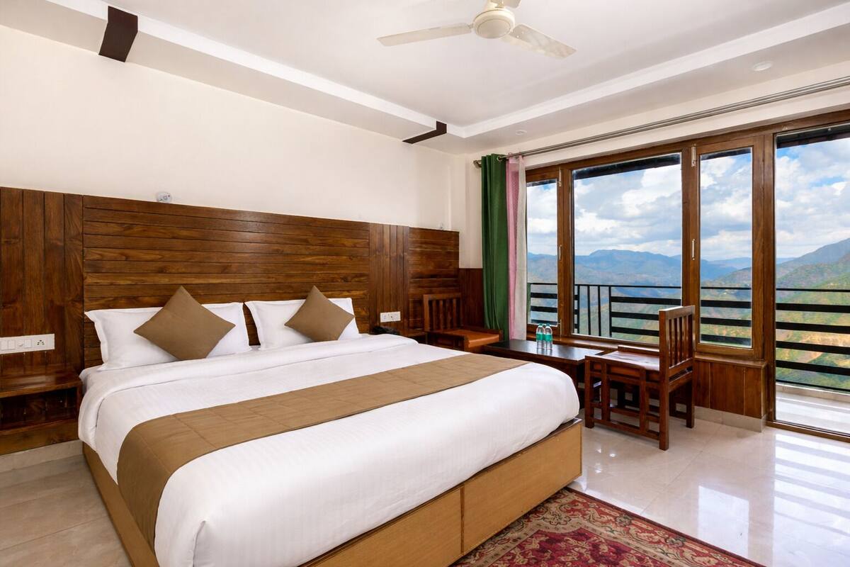 A hotel room with a large bed, a desk, a chair, a balcony with a mountain view, and a ceiling fan.