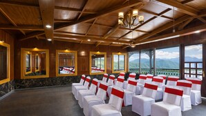 Une salle de conférence avec des rangées de chaises, des poutres en bois au plafond, de grandes fenêtres offrant une vue sur les montagnes et un lustre.
