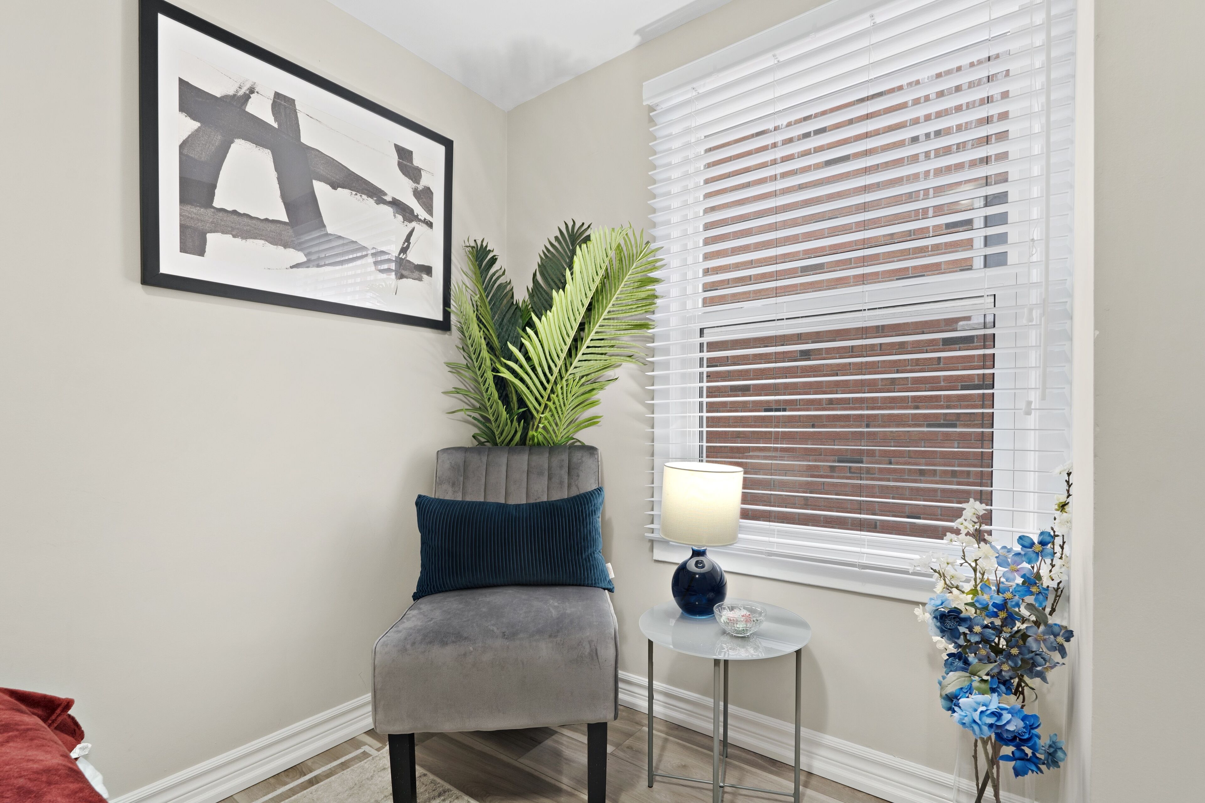A room with a grey upholstered chair, a small round table with a lamp, a framed abstract artwork, a potted plant, and a window with blinds.