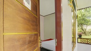 Eine Holztür mit gelbem Klebeband, ein Bett mit rotem und weißem Überwurf, ein Fenster mit Blick auf Grünflächen und ein kleiner Balkon mit einem Blumentopf.