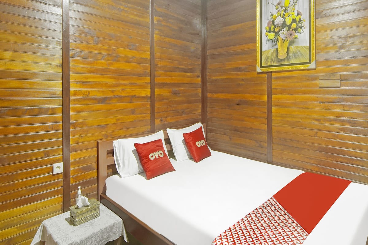 Ein Hotelzimmer mit einer holzverkleideten Wand, einem Bett mit weißer Bettwäsche und roten Kissen, einem kleinen Nachttisch mit Taschentuchbox und einer Flasche, sowie einem Blumengemälde an der Wand.