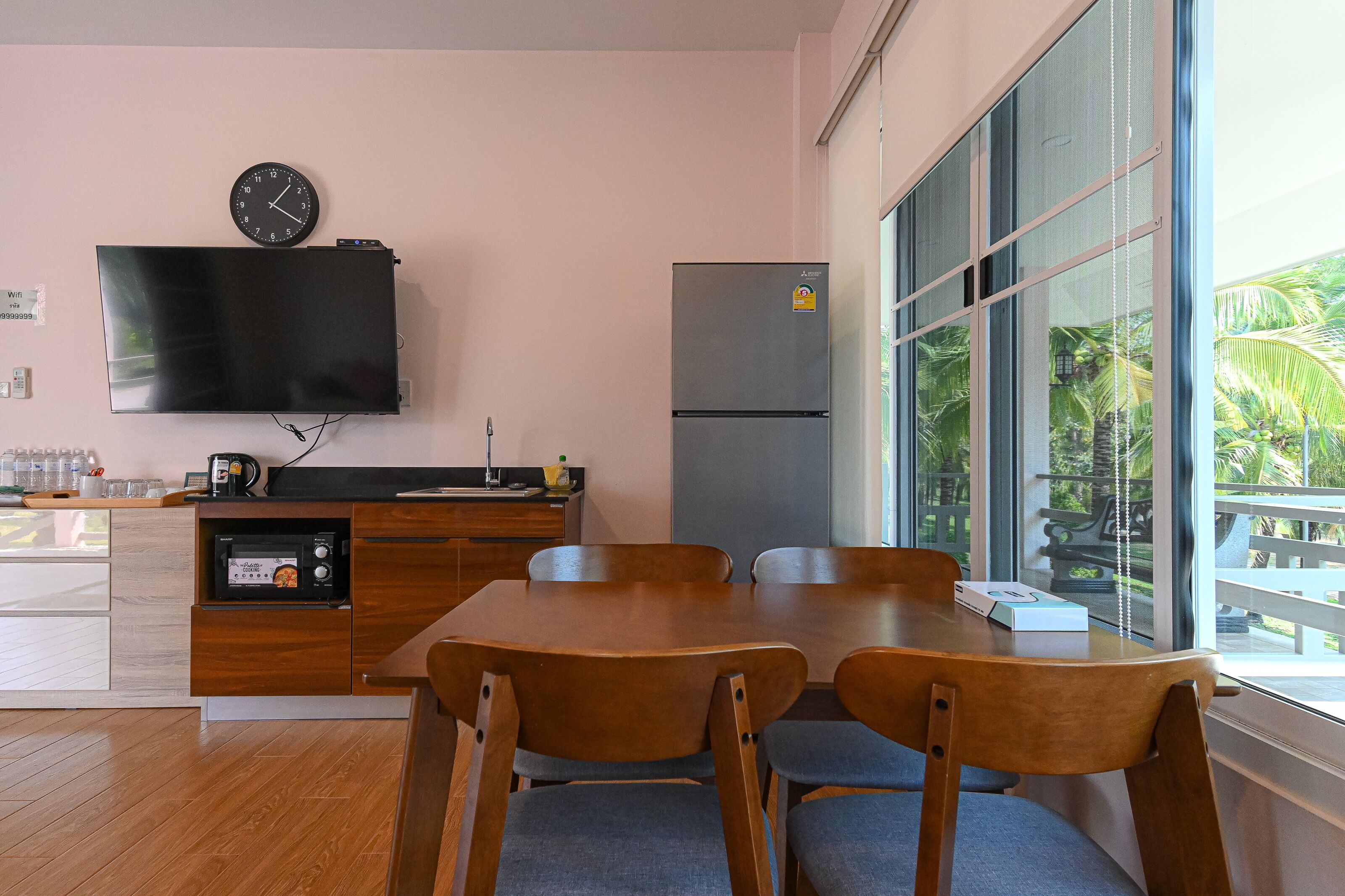 Una cocina moderna con televisor de pantalla plana, refrigerador, mesa de comedor de madera con sillas y una ventana con vista a palmeras.
