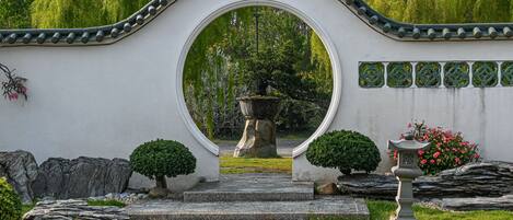A stone pathway leading to a traditional gate with a circular archway, surrounded by greenery and a stone lantern.