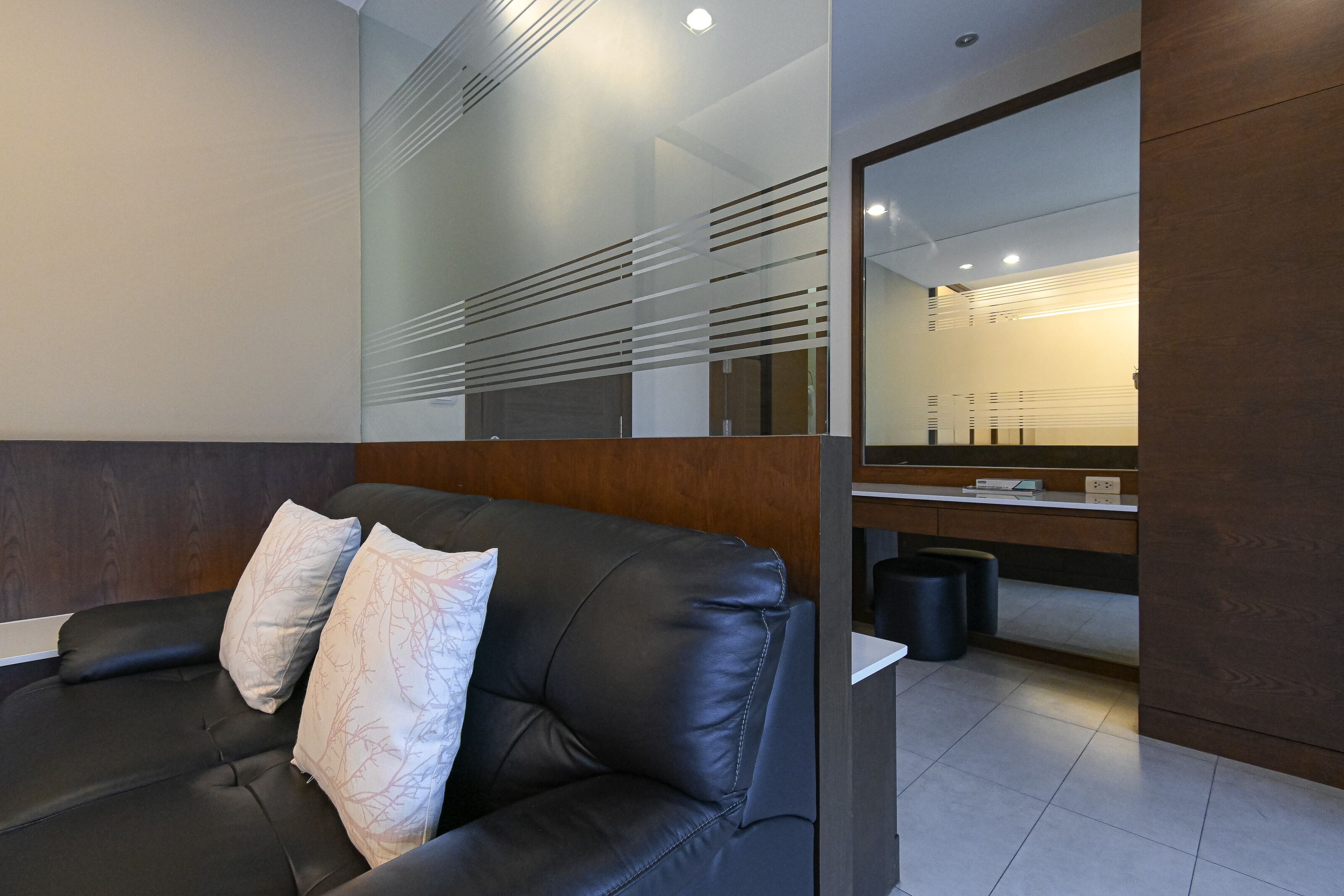 Une pièce moderne avec un canapé en cuir noir, un mur d’accent en bois et un grand miroir qui reflète une salle de bain bien éclairée.
