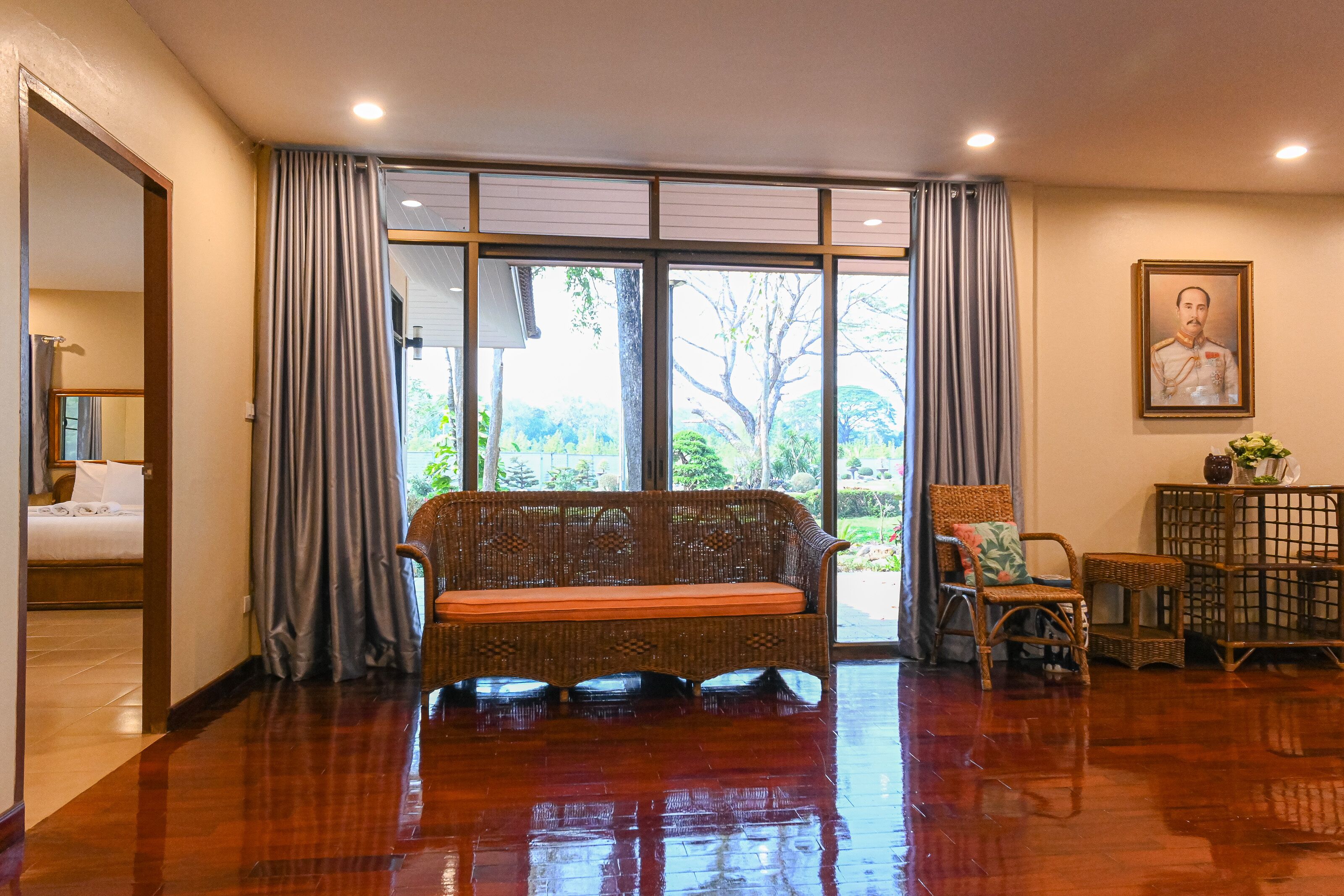 A room with a wooden floor, a wicker sofa with a cushion, a wicker chair, a small table with a vase, and a framed portrait on the wall.