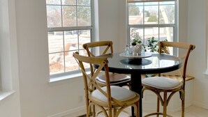 Un comedor con una mesa redonda, cuatro sillas de madera y una lámpara colgante negra. Se observan dos ventanas arqueadas con vistas al exterior.