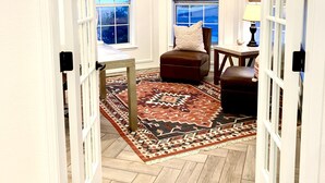 Una habitación con piso de madera, un tapete estampado, un sillón de cuero marrón, una mesita y una lámpara.