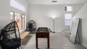 Una habitación con mesa de fútbol de mesa, una silla colgante, un banco y un ventanal con persianas.