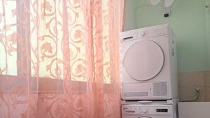 Ein Waschbereich mit übereinander gestapelter Waschmaschine und Trockner, einem Fenster mit leichtem Vorhang und einem an der Wand befestigten Ventilator.