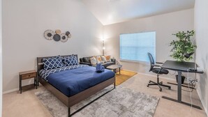 Une chambre à coucher comprenant un lit, des tables de chevet, un bureau, une chaise, un ventilateur de plafond, une fenêtre avec des stores et une plante.