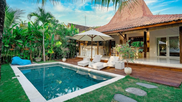 A swimming pool with a tiled deck, surrounded by lush greenery and a covered outdoor seating area with lounge chairs. - Villa Ketenangan (Ubud)