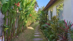 A narrow pathway flanked by lush tropical plants and a wooden fence, leading to a building with large windows. - Villa Ketenangan (Ubud)