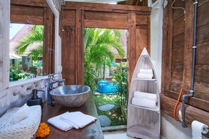 Un baño con un lavabo de piedra grande, estanterías de madera con toallas y vistas a un jardín tropical.