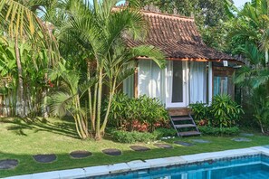 Una pequeña casa con techo de tejas, rodeada de abundante vegetación y una piscina.