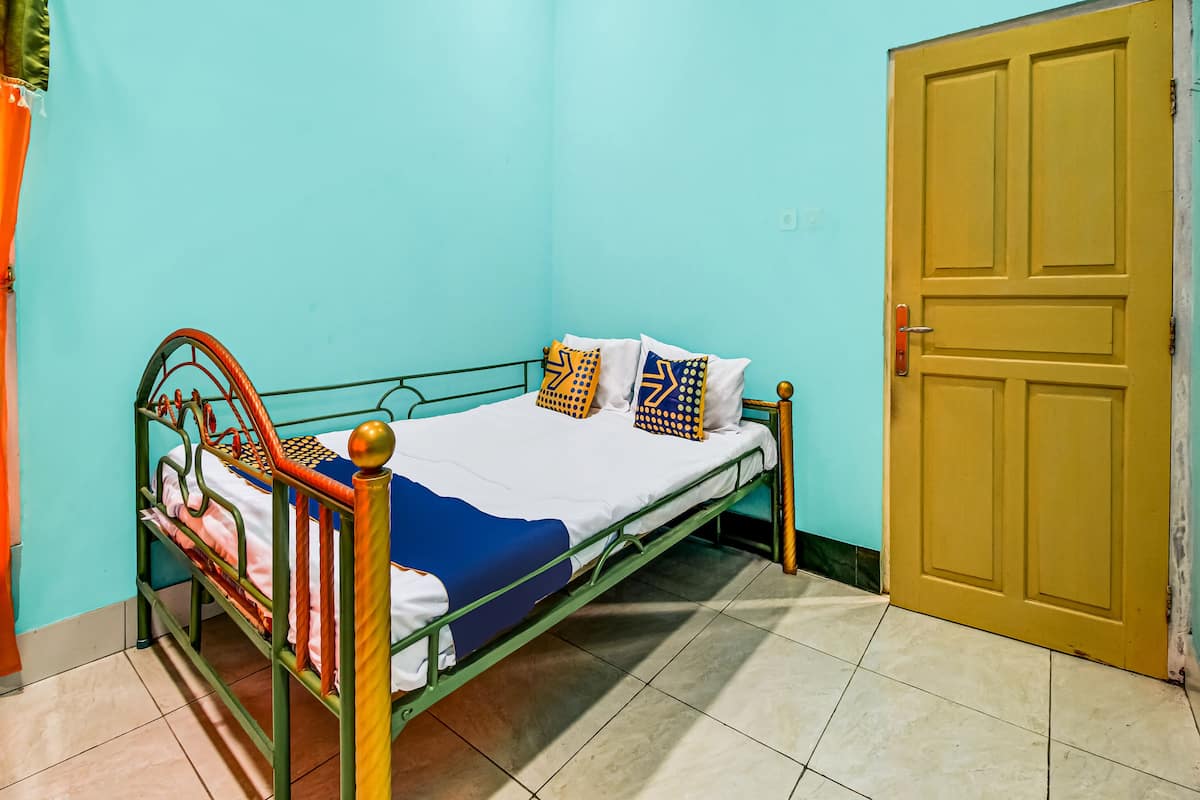 Une chambre avec un lit simple, un cadre en métal vert, du linge de lit blanc et des coussins à motifs. Une porte jaune est visible sur la droite.