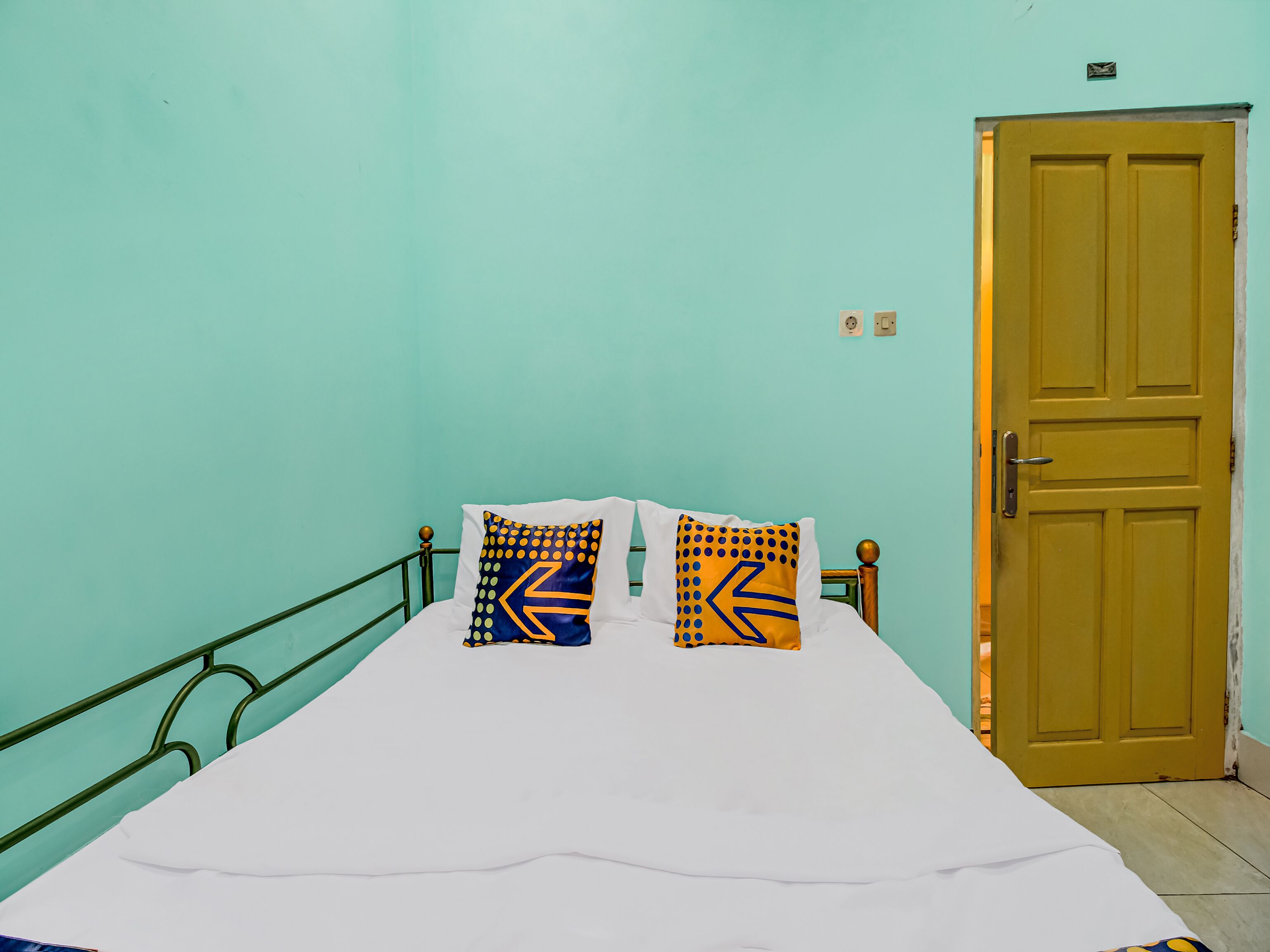 A bed with white bedding, two pillows with geometric patterns, a green metal headboard, and a wooden door.