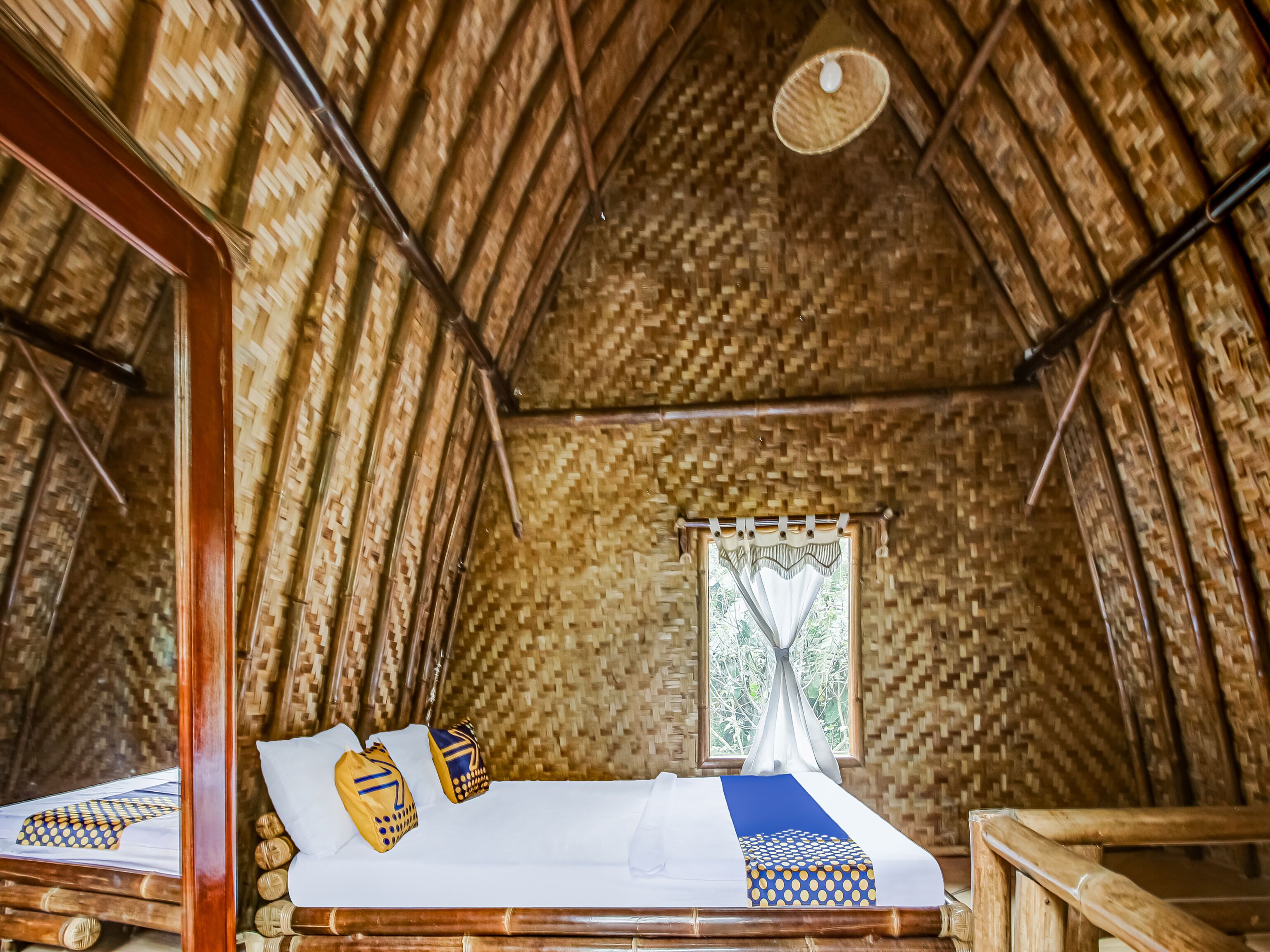 Una habitación con techo de paja, una cama con colcha blanca y azul, y una ventana con cortinas blancas.