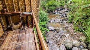 Habitación con piso de bambú, una pequeña mesa y un taburete, con vista a un arroyo rocoso y abundante vegetación.
